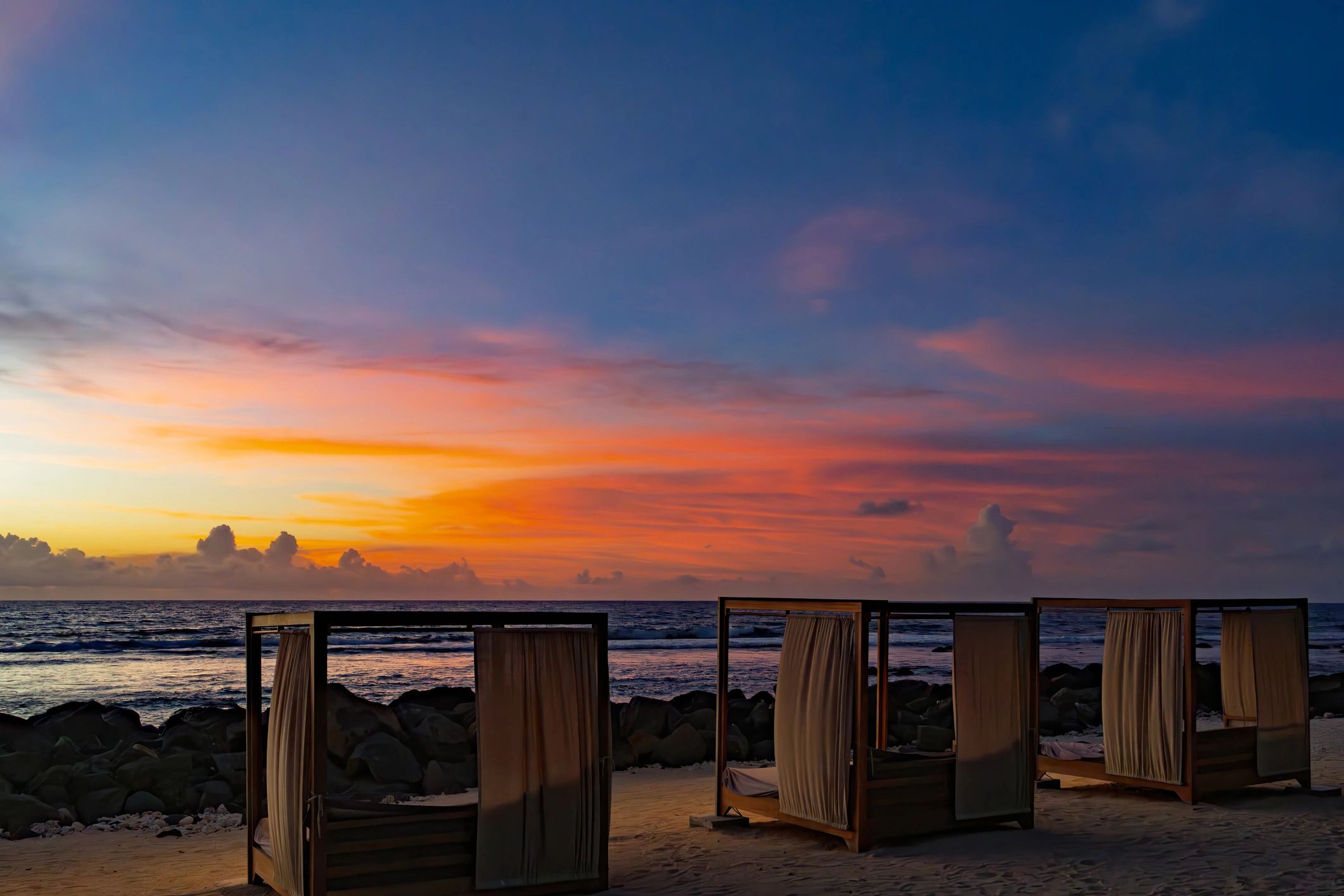 Wooden beach cabanas with flowing white curtains on sandy shore at vibrant sunset overlooking ocean in Thailand