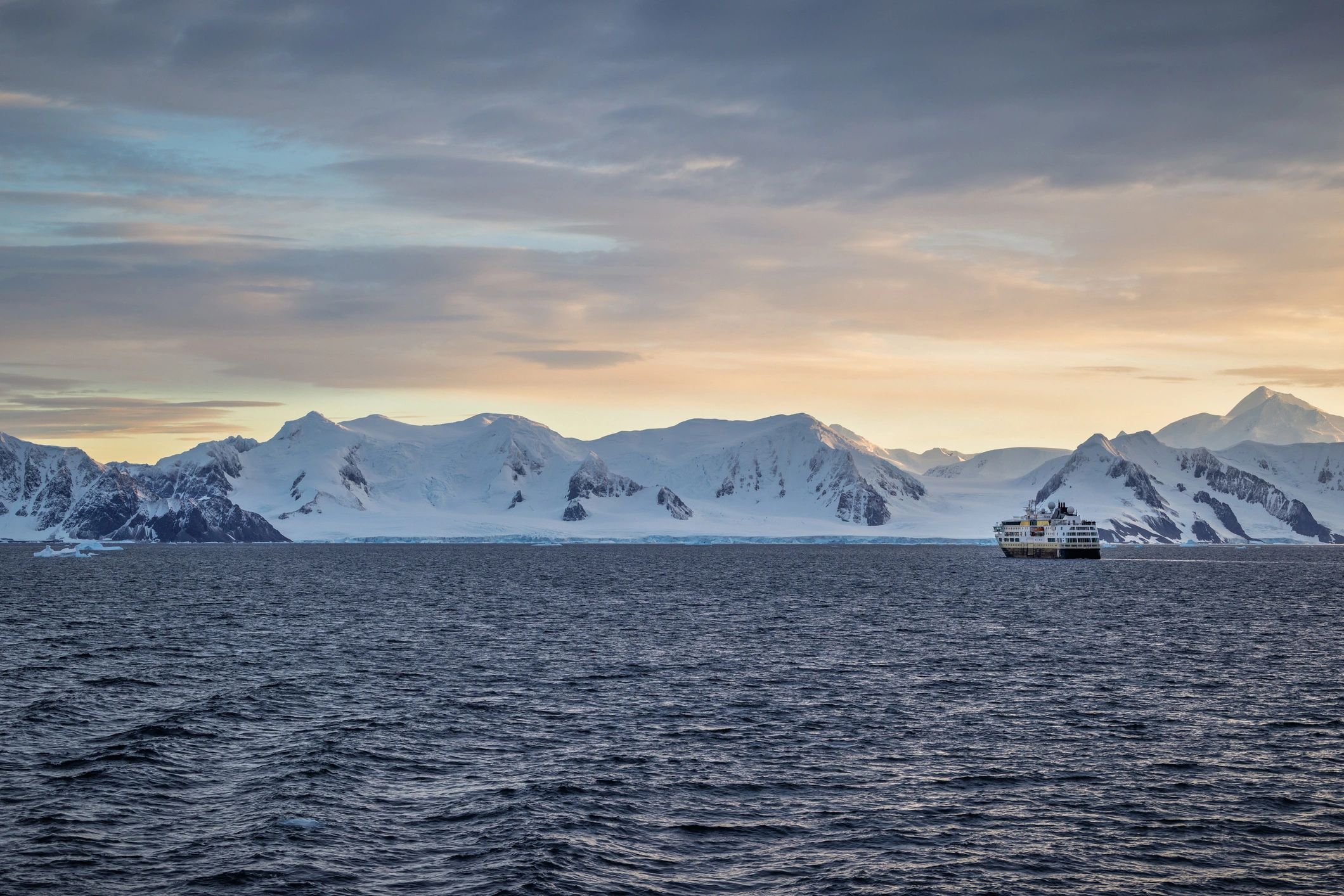 Expedition cruise ship sailing past snow-covered mountains and glaciers during sunset